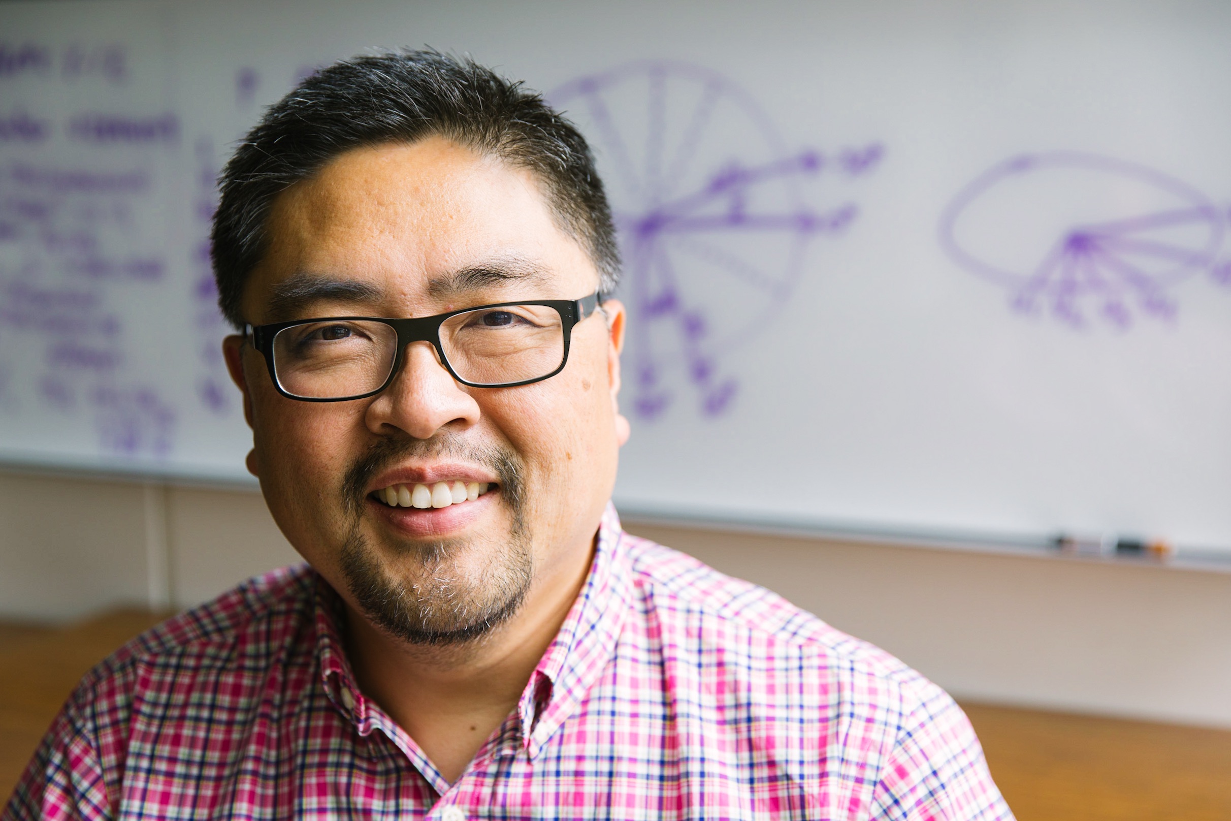 John Choe smiling, sitting in front of a whiteboard.